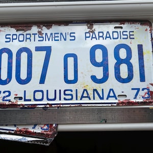 May include: A vintage Louisiana license plate, featuring the text "Sportsmen's Paradise" and the numbers "007 0 981". The plate is white with blue lettering, showing signs of wear and rust. The plate is from 1972-73.