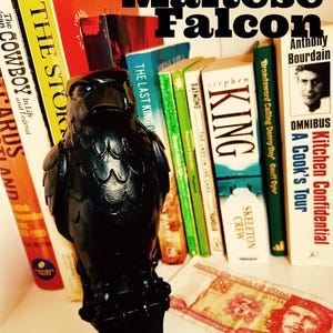 Puede incluir: Un sujetalibros de halcón negro en una estantería blanca con una variedad de libros, incluyendo "The Story of the Cowboy" y "Kitchen Confidential: A Cook's Tour" de Anthony Bourdain.