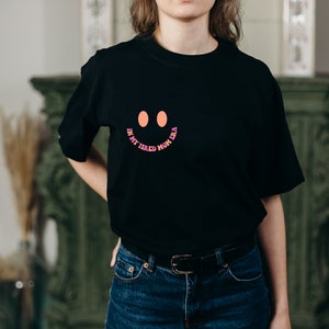 Pode incluir: Camiseta preta com um rosto sorridente rosa, laranja e roxo e o texto "In my tired mom era".