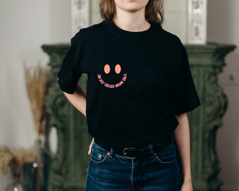 Pode incluir: Camiseta preta com um rosto sorridente rosa, laranja e roxo e o texto "In my tired mom era".