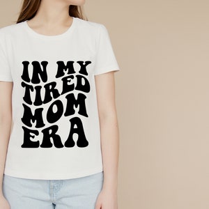Pode incluir: Camiseta branca com texto preto que diz "In My Tired Mom Era" em uma fonte retr&ocirc;.