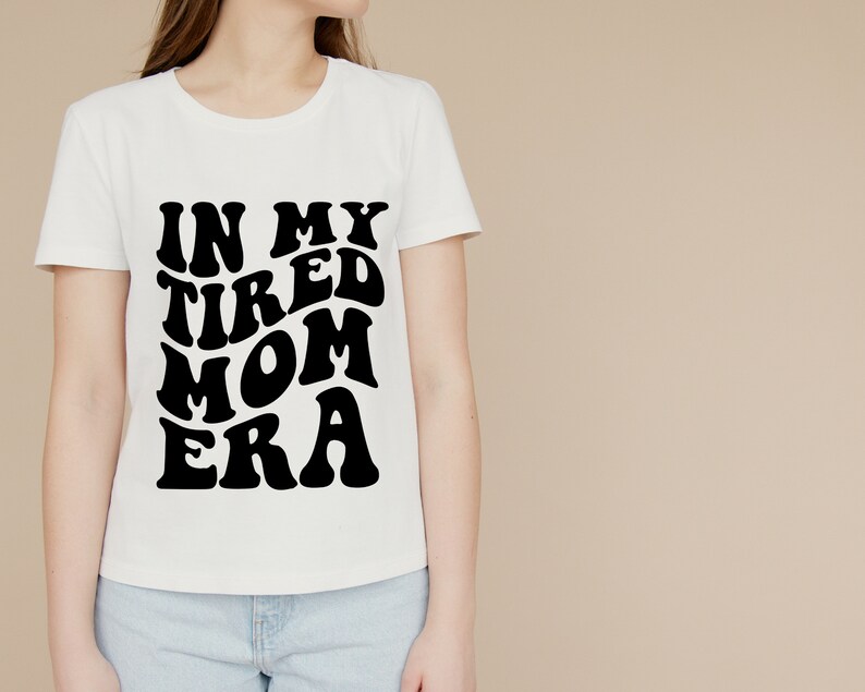 Pode incluir: Camiseta branca com texto preto que diz "In My Tired Mom Era" em uma fonte retr&ocirc;.