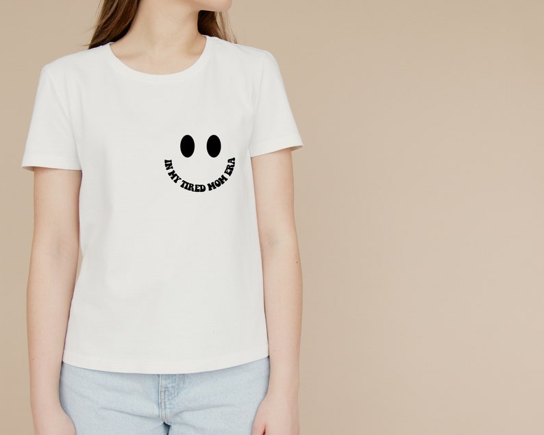 Pode incluir: Camiseta branca com um gr&aacute;fico de rosto sorridente preto e o texto "In my tired mom era".