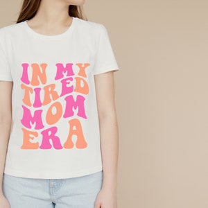 Pode incluir: Camiseta branca com um gr&aacute;fico rosa e laranja que diz "In My Tired Mom Era".