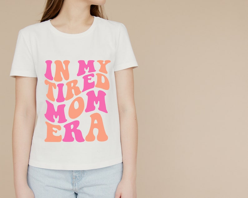 Pode incluir: Camiseta branca com um gr&aacute;fico rosa e laranja que diz "In My Tired Mom Era".