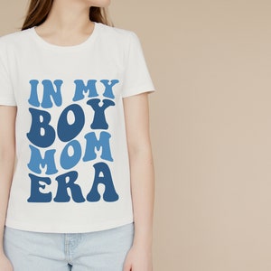 May include: White t-shirt with blue text that reads "In My Boy Mom Era" in a retro font.