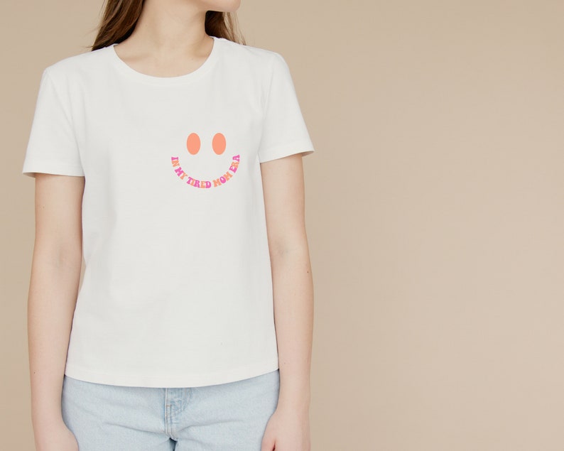 Pode incluir: Camiseta branca com um rosto sorridente rosa e laranja e o texto "In my tired, home era".