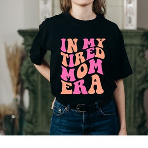 Pode incluir: Uma camiseta preta com o texto "In My Tired Mom Era" em letras rosa e laranja. As letras est&atilde;o em uma fonte retro ondulada.