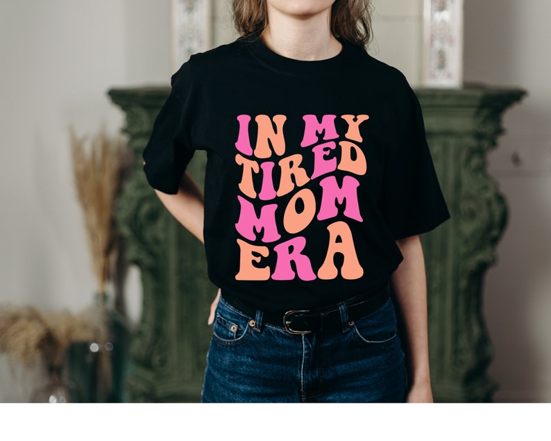 Pode incluir: Uma camiseta preta com o texto "In My Tired Mom Era" em letras rosa e laranja. As letras est&atilde;o em uma fonte retro ondulada.