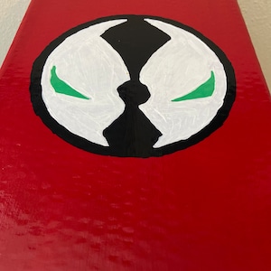 May include: A red rectangular object with a Spawn logo painted on the top. The logo is a white, black, and green circular design. The background is a solid, glossy red.