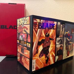 May include: A rectangular black comic book storage box featuring a Marvel Blade comic cover graphic on the side, showing the character Blade in action. A tall red box with the word BLADE printed in black is positioned behind the storage container.
