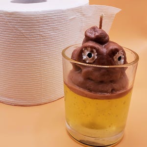 May include: A brown poop-shaped candle with googly eyes sits in a clear glass jar filled with a yellow liquid. A roll of white toilet paper is in the background.