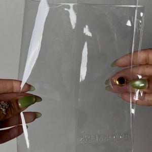 May include: Clear plastic bookmark holder with a gold-colored snap closure. The holder is rectangular and transparent, allowing visibility of the bookmark inside. The word "BOOKMARK" is embossed on the bottom.