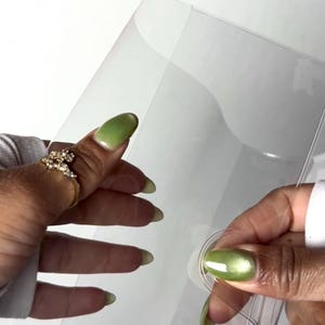 May include: Clear plastic book cover with a circular closure. The cover is held by a person with green nail polish and a gold ring. The text "BOOKMARK" is visible on the cover.