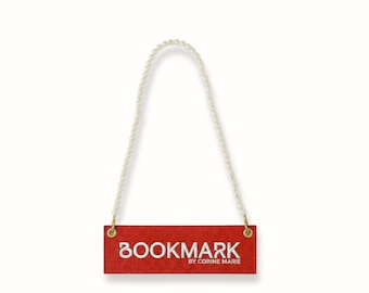 May include: A red bookmark with the word "BOOKMARK" in white script. The bookmark has a pearl beaded handle and the text "BY CORINE MARIE" below the word "BOOKMARK".