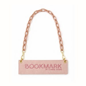 Puede incluir: Un bolso rectangular rosa con la palabra "BOOKMARK" bordada y una cadena como asa. El bolso tiene cierres dorados y el texto "BY CORINE MARIE" debajo de la palabra "BOOKMARK".