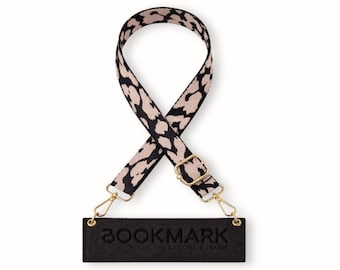 May include: A black and tan leopard print strap with gold-colored hardware and a black rectangular tag that reads "BOOKMARK by CORINE MARIE". The strap is designed to be used with a bag or purse.