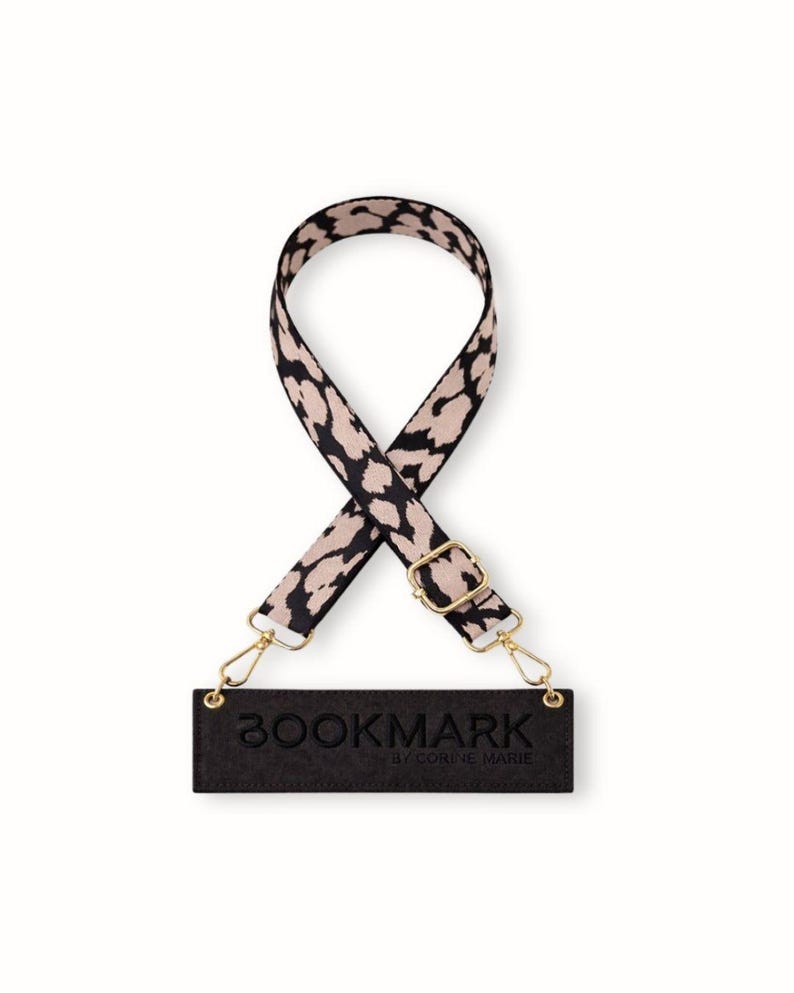 May include: A black and beige leopard print strap with gold-coloured metal fixings and a black rectangular tag that reads "BOOKMARK by CORINE MARIE". The strap is for a bag or purse.