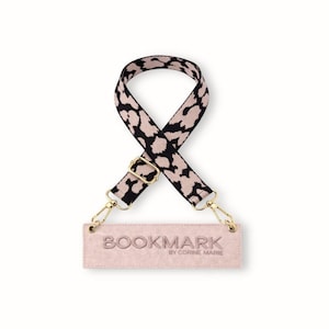 May include: A stylish purse strap with a black and tan leopard print pattern. The strap has gold-colored metal clasps and a rectangular, blush-colored tag that reads "BOOKMARK BY CORINE MARIE."