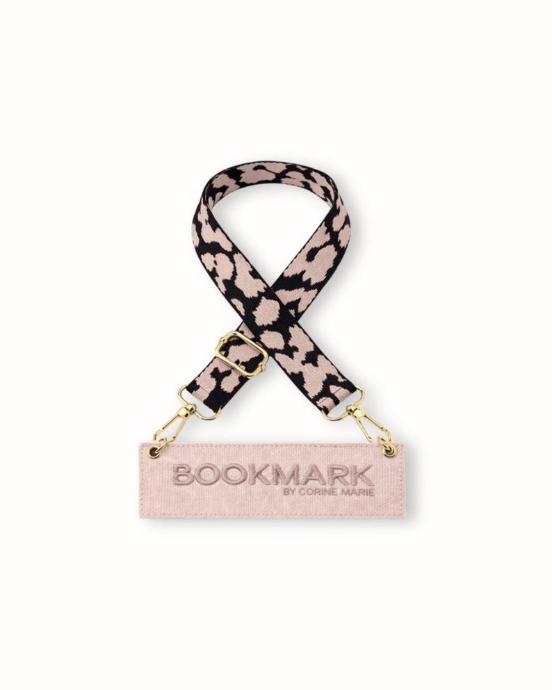 May include: A patterned handbag strap featuring a black and beige leopard print. It has gold-tone metal clasps and a blush-pink tag with the text "BOOKMARK BY CORINE MARIE".