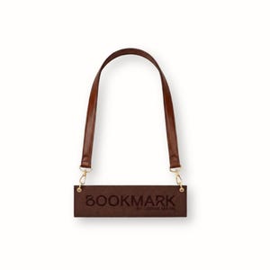 Puede incluir: Un bolso rectangular marrón con un asa larga y curvada. El bolso tiene herrajes dorados y la palabra "BOOKMARK" está grabada en relieve en la parte delantera, junto con el texto "BY COPINE MARIE".
