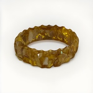 Puede incluir: Un anillo circular con una base de resina translúcida de color ámbar incrustada con numerosos fragmentos pequeños e irregulares de color blanco hueso y marrón claro. La superficie exterior de la banda tiene un aspecto texturizado y facetado.