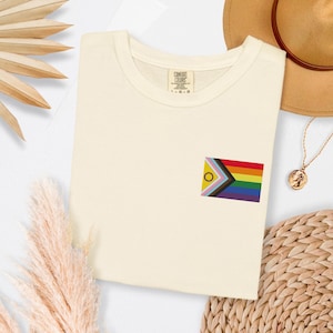 Puede incluir: Camiseta color crema con una pequeña bandera bordada con un arcoíris y otros colores. La camiseta tiene cuello redondo y una etiqueta que dice "COMFORT COLORS". Un collar dorado y un sombrero marrón son visibles en el fondo.