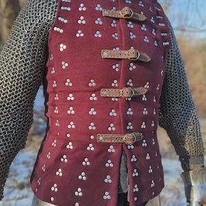May include: A burgundy medieval gambeson vest with silver rivet detailing and brown leather straps. The vest features a high collar and is worn with chainmail armor. The design includes three leather straps with buckles.