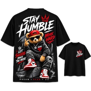 Peut inclure: Un t-shirt noir avec un grand motif d'ours en peluche portant une casquette rouge, des lunettes de soleil et des baskets, avec le texte STAY HUMBLE, WORK HARD, LET SUCCESS MAKE THE NOISE, NEVER GIVE UP et DREAM PLAN DO. Une version plus petite montre une couronne et LEGEND.