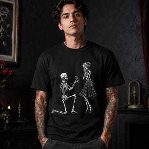 May include: A black short-sleeved t-shirt featuring a white graphic print of two skeletons. One skeleton is kneeling on one knee and holding an open ring box towards the other skeleton, who is standing and wearing a skirt and high-heeled shoes.