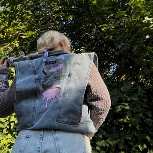 Puede incluir: Un chaleco vaquero con bordados en morado y negro. El bordado incluye las palabras "Free" y "Wild" y un diseño de labios morados que gotean.