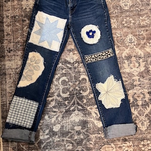 May include: Dark wash denim jeans with various fabric patches. Patches include a light blue star, a white and blue floral design, a gingham pattern, a leopard print, and a white leaf design. The jeans have cuffed hems.