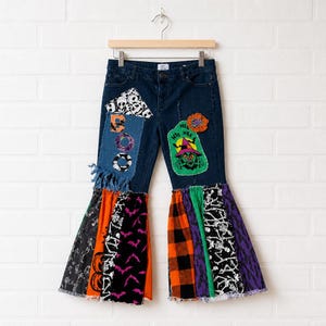 May include: Dark blue denim jeans featuring a flared leg design with patchwork accents. The pants include various Halloween-themed fabric patches, including skeletons, bats, pumpkins, and a witch graphic with the text "HEE HEE" and "BOO".