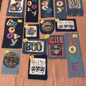 May include: A collection of fourteen rectangular denim fabric patches featuring various graphic designs, including cats, a rooster, an eye, and text such as Groovy, Peace, Joy, Boo, Free 2 B Me, and Love, each labeled with a small yellow numbered sticker.