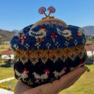 May include: A knitted coin purse with a blue background featuring white chickens and red mushrooms. The purse has a gold clasp with two purple gemstone accents. The bottom of the purse is black with white chickens. The purse is held in a hand.