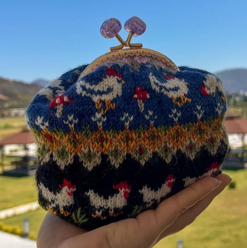 May include: A knitted coin purse with a blue background featuring white chickens and red mushrooms. The purse has a gold clasp with two purple gemstone accents. The bottom of the purse is black with white chickens. The purse is held in a hand.