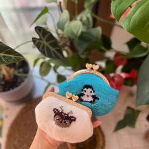 May include: Two knitted coin purses. One is light blue with a penguin design, the other is off-white with a deer design. Both have gold-colored heart-shaped clasps and tan trim.