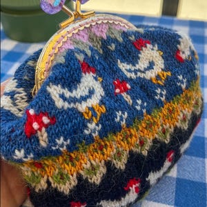 May include: A knitted coin purse with a gold clasp and a decorative top. The purse features a blue background with white chickens and red mushrooms. The top of the purse has a pink and purple floral design.