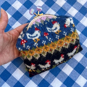 May include: A knitted coin purse with a clasp closure. The purse features a blue background with white chickens and red mushrooms. The top of the purse has a pink and purple floral design. The clasp is gold-colored with purple accents.