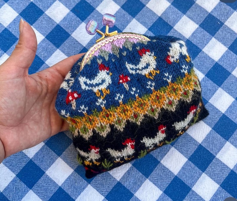 May include: A knitted coin purse with a clasp closure. The purse features a blue background with white chickens and red mushrooms. The top of the purse has a pink and purple floral design. The clasp is gold-colored with purple accents.