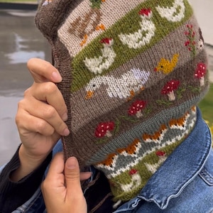 May include: A hand-knitted hat and scarf set featuring a whimsical farm animal design. The hat showcases ducks, chickens, and mushrooms in shades of brown, green, and white. The scarf has a matching pattern. A cozy, handmade accessory.