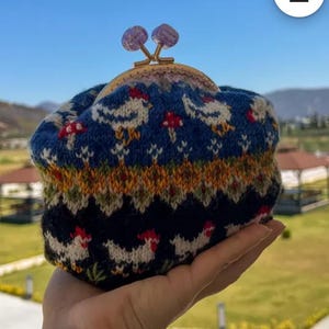 May include: A knitted coin purse with a blue background featuring white chickens and red mushrooms. The purse has a gold-colored clasp and is topped with two purple decorative beads. The purse is held in a hand.