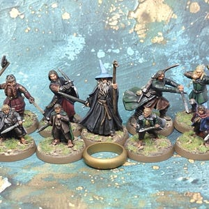 May include: A collection of miniature figurines depicting characters from a fantasy setting. The figures are painted with detailed clothing and weaponry, and are mounted on circular bases. A gold ring is in the foreground.