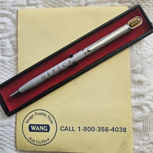 May include: A silver pen with the word "OFFICE" printed on it. The pen is in a black box with a red interior. The box has a gold sticker with the word "WANG" on it. The pen is on a yellow card with the text "Image Enable Now WANG Ask Us How CALL 1-800-356-4038".