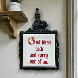 May include: Black metal wall plaque with a decorative frame and a church-shaped top. The white tile center features the text "God bless each and every one of us" in red script.