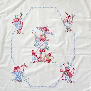 May include: A white fabric cloth featuring red, blue, and green embroidered circus-themed illustrations. The design includes clowns, a monkey, a duck, and an elephant arranged around a central carousel figure, all enclosed by a blue geometric border.
