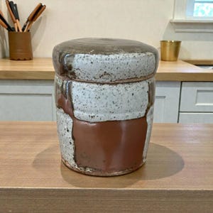 May include: A ceramic jar with a lid. The jar has a speckled white and brown exterior with a band of solid brown around the middle. The lid is a darker brown. The jar is sitting on a wooden surface.