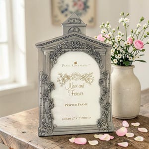 May include: A pewter photo frame with a floral design and the words "Now and Forever." The frame holds a 5" x 7" photo. The frame is gray and has a decorative top. A vase of flowers is next to the frame.
