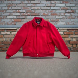May include: A vibrant red jacket with a collared neckline, two front pockets, and a ribbed hem and cuffs. The jacket has a button closure and a small tag on the collar. The background is a brick wall.