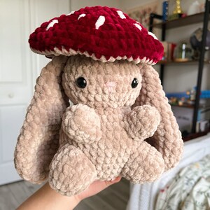 May include: A plush bunny toy with a red mushroom cap with white spots. The bunny is a light brown color with black eyes and a pink nose. The mushroom cap is a deep red color. The toy is soft and cuddly.
